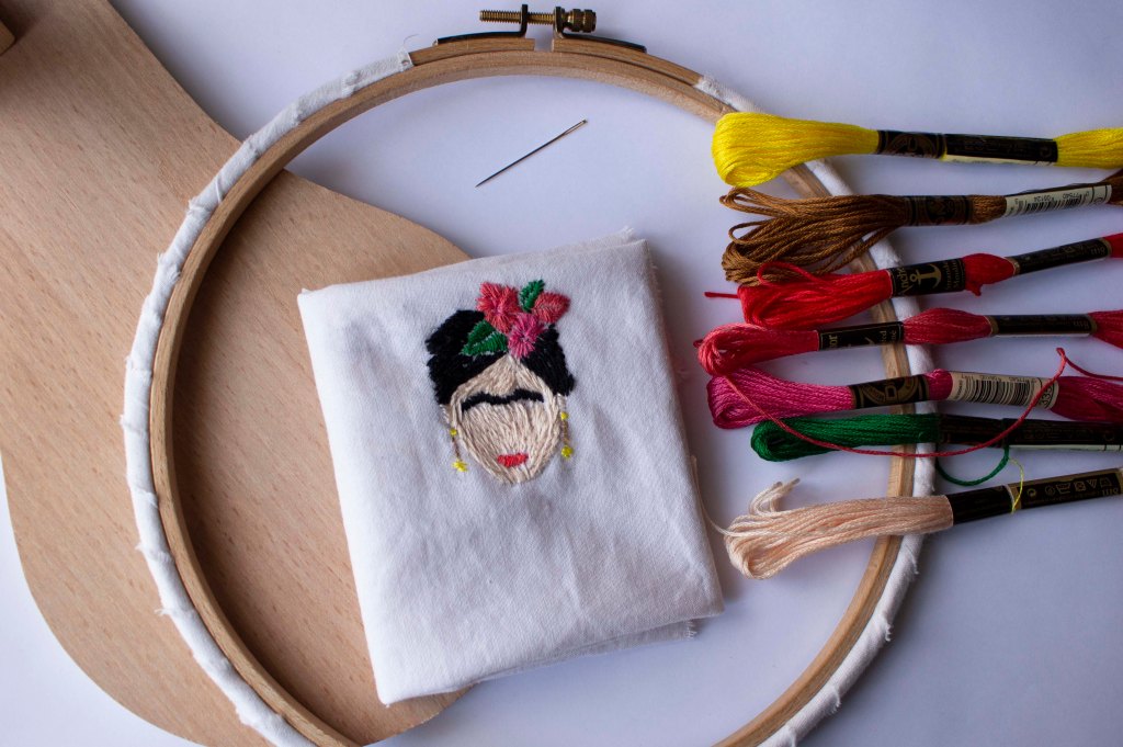Frida Kahlo Face embroidery onto white cloth surrounded by embroidery threads and sewing needle.